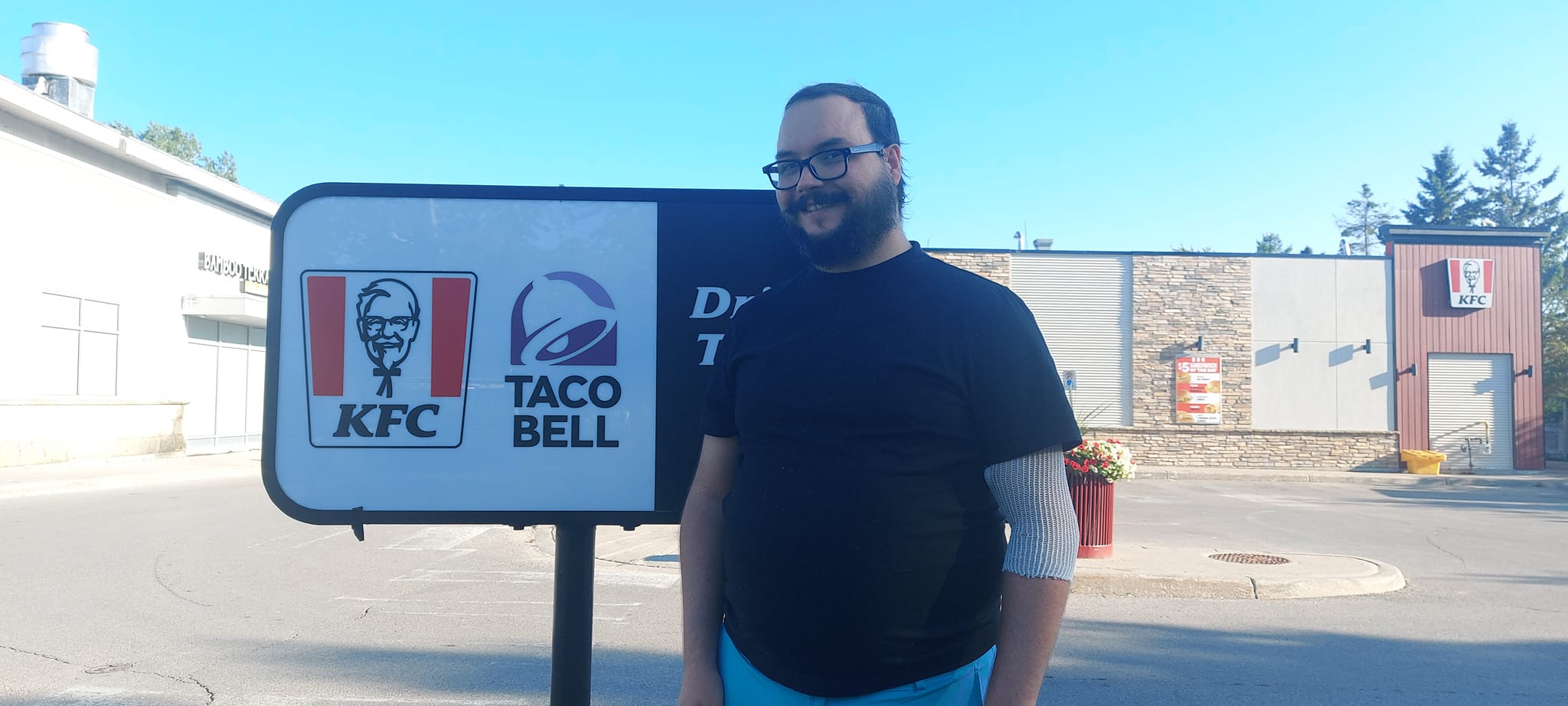 A man short hair and bushy facial hair stands in front of a KFC/Taco Bell sign